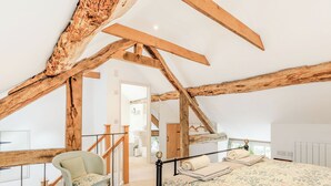 Ein Schlafzimmer mit freiliegenden Holzbalken, einem Bett mit floralem Bettzeug, einem Sessel und einer Treppe, die zu einem anderen Raum führt.