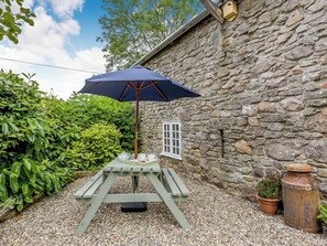 A rustic stone wall, a wooden table with a blue umbrella, and a window with white shutters.