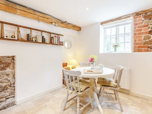 A cozy dining area with a round table, white chairs, a wooden ceiling, a brick wall, and a window with flowers.