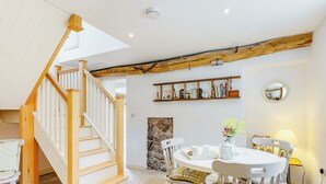Ein geräumiges Zimmer mit einer Holztreppe, einem Essbereich mit weißen Stühlen, einem Steinkamin und einem Bücherregal.