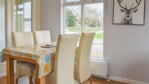 Ein Essbereich mit Tisch, der mit einer gemusterten Tischdecke bedeckt ist, vier Stühle, einem Fenster mit Blick auf Grünflächen und einem gerahmten Bild an der Wand.