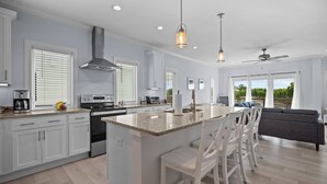 A modern kitchen with white cabinetry, a central island with bar stools, and a dining area with a gray sofa. - Modern Single Family Home 5 minute walk to the Beach! Linens Included! (Carolina Beach)