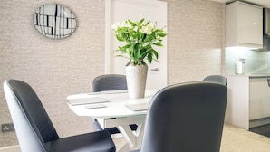 A modern dining area with a glass table, black chairs, a potted plant, and a kitchen in the background.