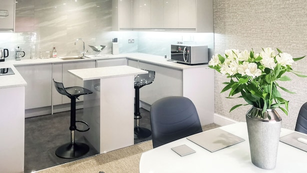 A modern kitchen with white cabinets, a microwave, and a coffee maker. There are two bar stools and a dining table with a vase of flowers.