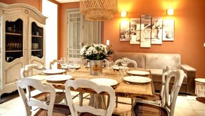 Zona pranzo con un tavolo di legno apparecchiato per sei persone, circondato da sedie bianche. Sul lato sinistro, un armadietto bianco con ante in vetro, e sopra il tavolo, un lampadario in vimini.