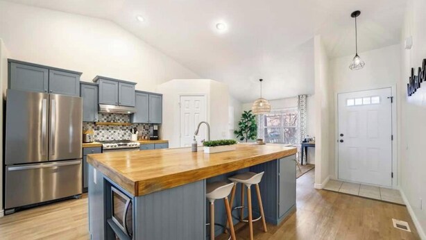 Een moderne keuken met grijze kastjes, roestvrijstalen apparatuur, een houten eiland met krukken en een uitzicht op de stad door grote ramen.