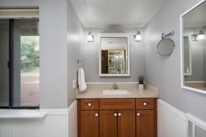 Un baño con tocador de madera, un lavamanos y un espejo. Se ve una ventana con vistas al exterior.