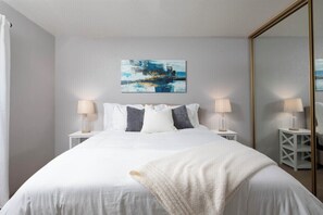Un dormitorio ordenado con una cama grande, dos mesitas de noche con lámparas, un cuadro abstracto en la pared y un espejo de cuerpo entero.