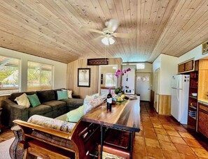 A cozy living room with a wooden ceiling, a sofa, a coffee table, and a kitchen area with a refrigerator and cabinets.