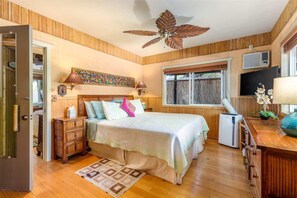 A bedroom with a bed, wooden walls, a ceiling fan, a window, a desk, and a TV.