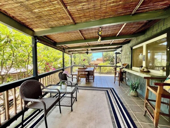 A covered outdoor seating area with tables and chairs, a bar, and a view of greenery.
