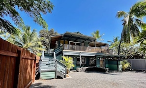 A two-story building with a balcony, surrounded by palm trees and a wooden fence.