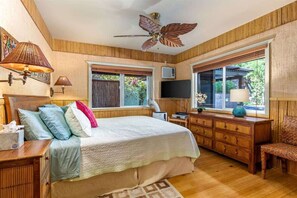 A bedroom with a bed, wooden headboard, bedside lamps, a ceiling fan, a TV, a wooden dresser, a chair, and large windows with a view of greenery.