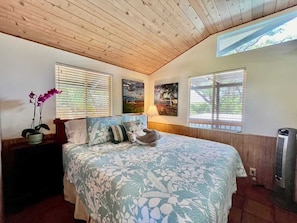 Une chambre avec un plafond en bois, un lit recouvert d’une literie à motifs floraux, une table de chevet ornée d’une plante en pot et un radiateur fixé au mur.