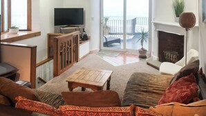 A living room with a TV, sofa, and a view of the sea through a glass door. - Reverse level cottage in Newlyn stunning sea views from the roof top balcony. (Newlyn)