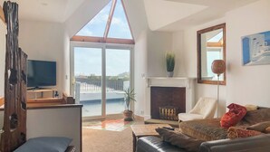 A living room with a TV, a fireplace, a sofa, and a view of a dock. - Reverse level cottage in Newlyn stunning sea views from the roof top balcony. (Newlyn)