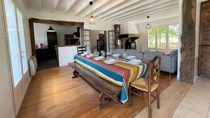 A dining area with a wooden table set for four, a bench, and a colorful tablecloth. There is a kitchen in the background with a stove and a clock.