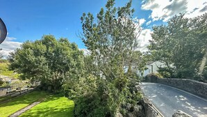 Un pont de pierre mène à un bâtiment blanc doté d’un toit en pente, entouré d’une végétation luxuriante et surplombé d’un ciel bleu clair.