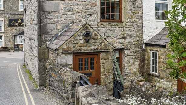 Un bâtiment en pierre avec une porte et une fenêtre en bois, situé à côté d’une route où se trouve un panneau indiquant « The Crow ».