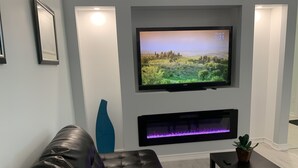 A modern living room with a leather sofa, a flat-screen TV mounted above a fireplace, and a small table with a plant. - Waterfront Cottage on the Upper Rideau Lake (Westport)