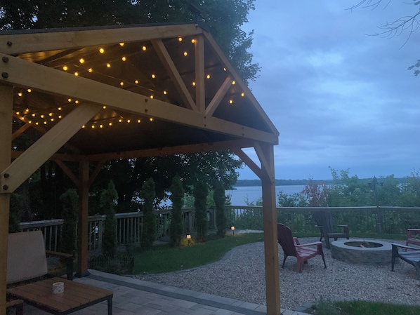 A wooden pergola with string lights, outdoor seating, and a view of a lake.