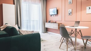 Un saló modern amb un sofà verd, una taula de dinar amb cadires, un televisor fixat a la paret i un petit escriptori amb un ordinador portàtil.