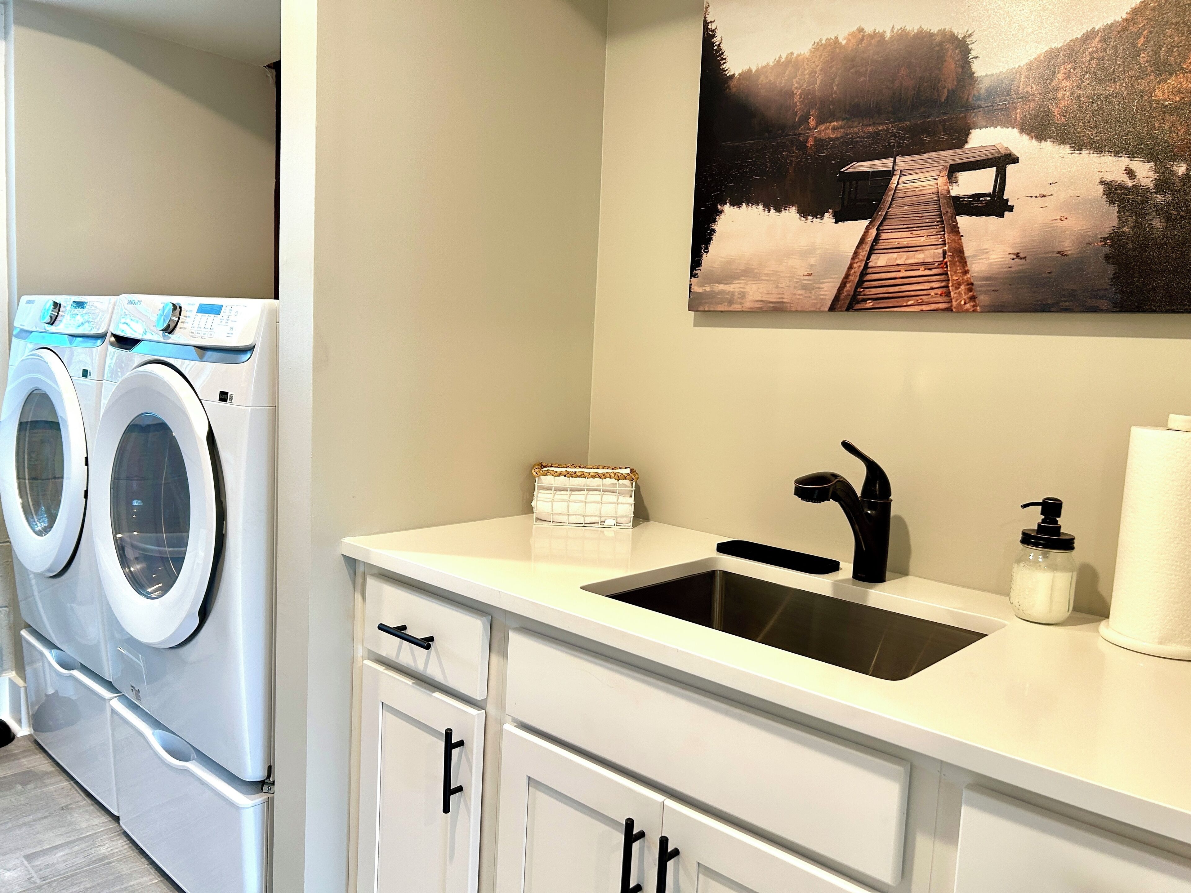 A laundry room with two front-loading washing machines, a white countertop with a black faucet, a soap dispenser, and a paper towel holder.