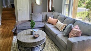 A living room with a grey sectional sofa, a round coffee table, and a view of a garden through large windows. - Lake Front Haven with Dual Living Rooms, Hot Tub (Mt. Juliet)