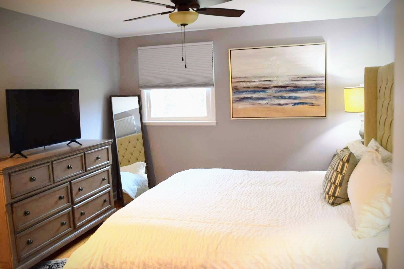 A bedroom with a bed, a dresser, a television, a ceiling fan, a window with blinds, and a painting on the wall.