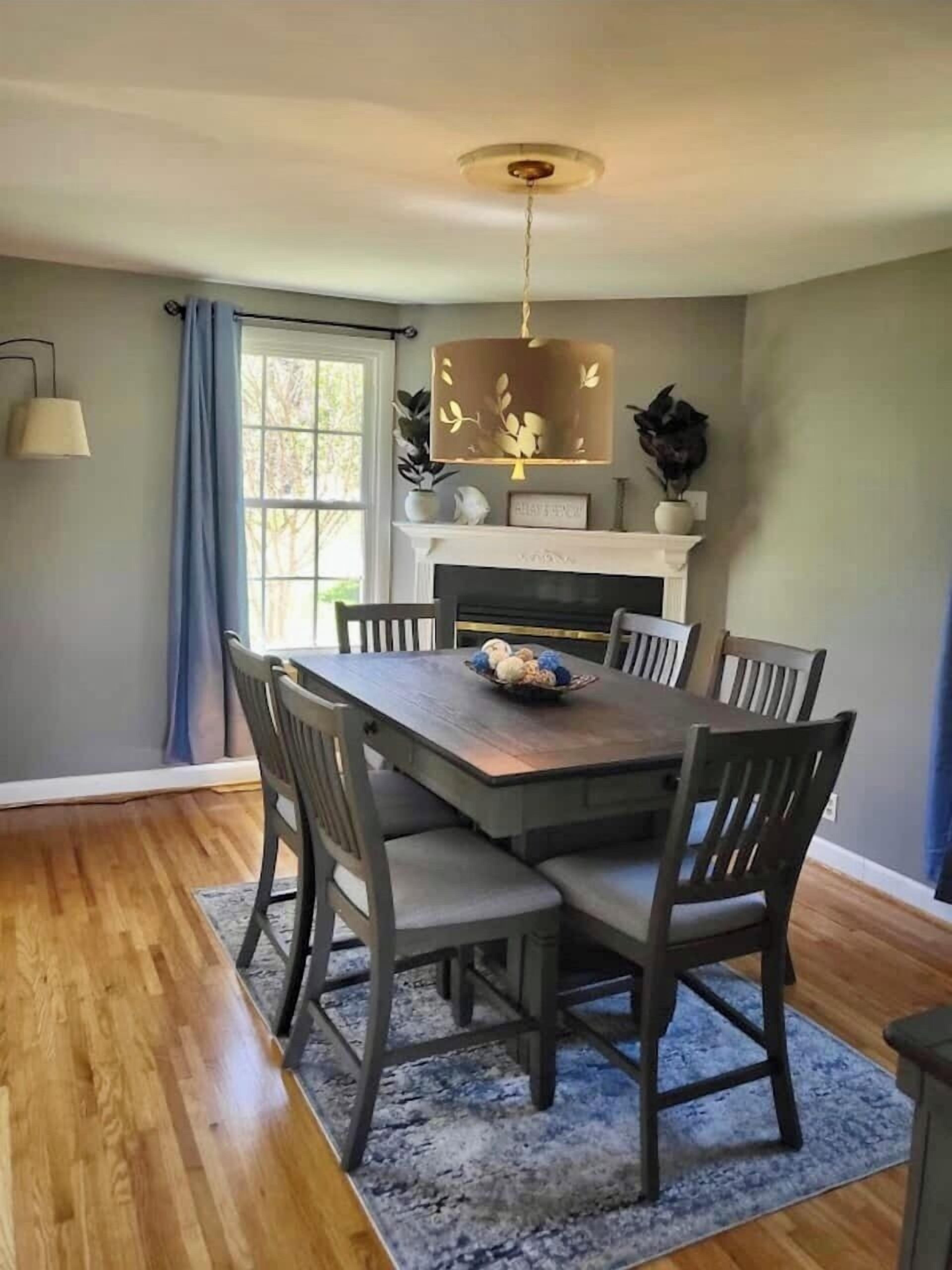 Comedor con mesa de madera, seis sillas, candelabro, chimenea y ventana con cortinas.