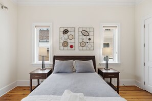 A bedroom with a bed, two nightstands, a window, and a wall art piece. - Heart of King St · Charming Condo· Private Balcony (Charleston)