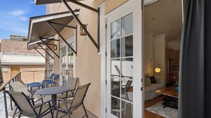 Um pátio externo com mesa e cadeiras, uma porta de vidro deslizante que dá para um espaço de estar interno, e vista dos edifícios ao redor.