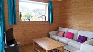 Ein gemütliches Zimmer mit Holzvertäfelung, einem weißen Sofa mit bunten Kissen, einem Holztisch und einem Fernseher auf einem schwarzen Lowboard.