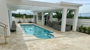 A covered outdoor swimming pool with a tiled deck, lounge chairs, and a view of the surrounding greenery.