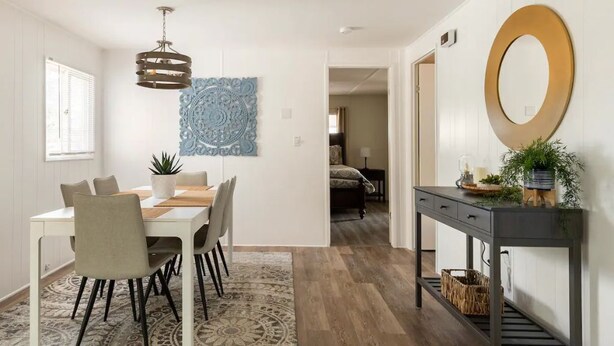 A dining area with a table, chairs, a patterned rug, a pendant light, and a console table with a mirror and plants.