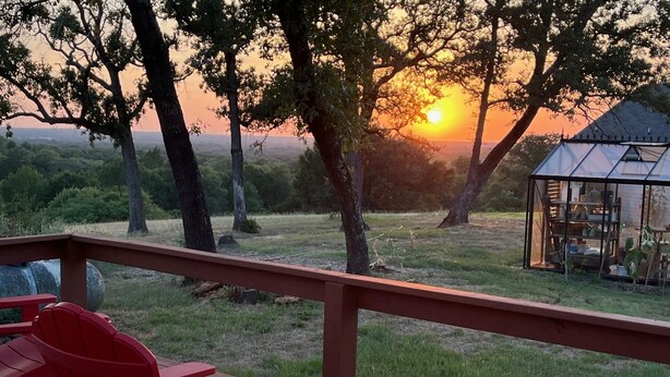 A wooden deck with red Adirondack chairs, a glass greenhouse, and a view of the sunset over a grassy landscape with trees. - Hilltop Cottage Sunset Views at The Farm on a Hill (Caldwell)
