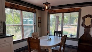 A dining area with a round table and wooden chairs, large windows with blinds, and a wooden ceiling beam. - Hilltop Cottage Sunset Views at The Farm on a Hill (Caldwell)