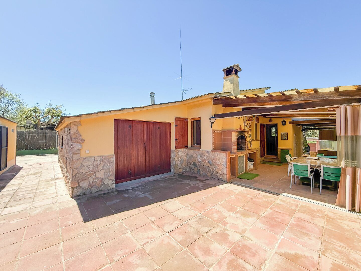 Une terrasse avec un sol en pierre, une porte en bois et un espace barbecue comprenant une table et des chaises.