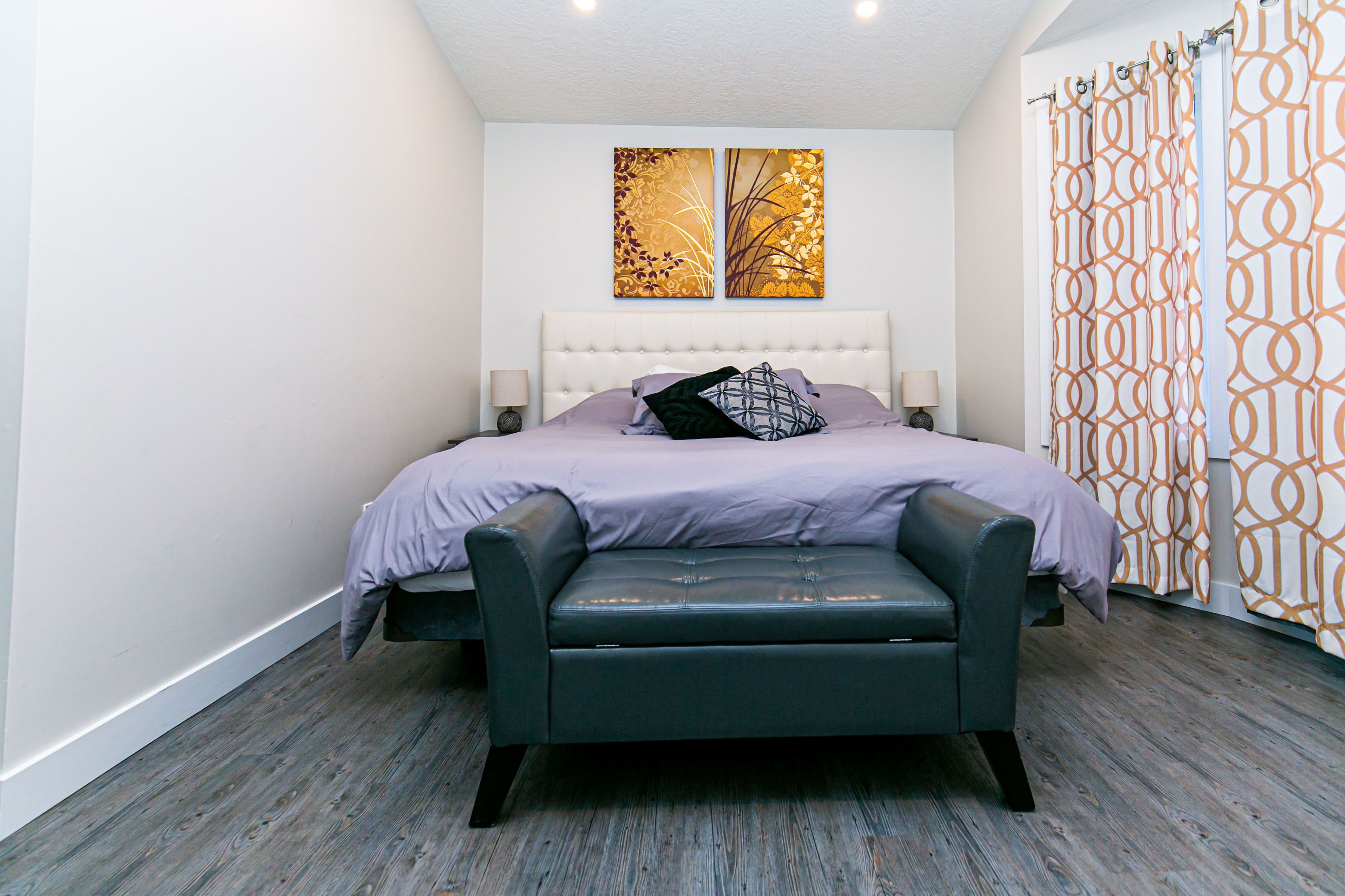 A modern bedroom with a tufted headboard, a bed with a purple bedspread, a black leather bench, and two wall-mounted artworks.
