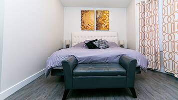 A modern bedroom with a tufted headboard, a bed with a purple bedspread, a black leather bench, and two wall-mounted artworks.