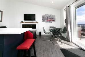 A modern kitchen with a flat-screen TV, a dining area with a glass table and chairs, a red bench, and a large window with a view.
