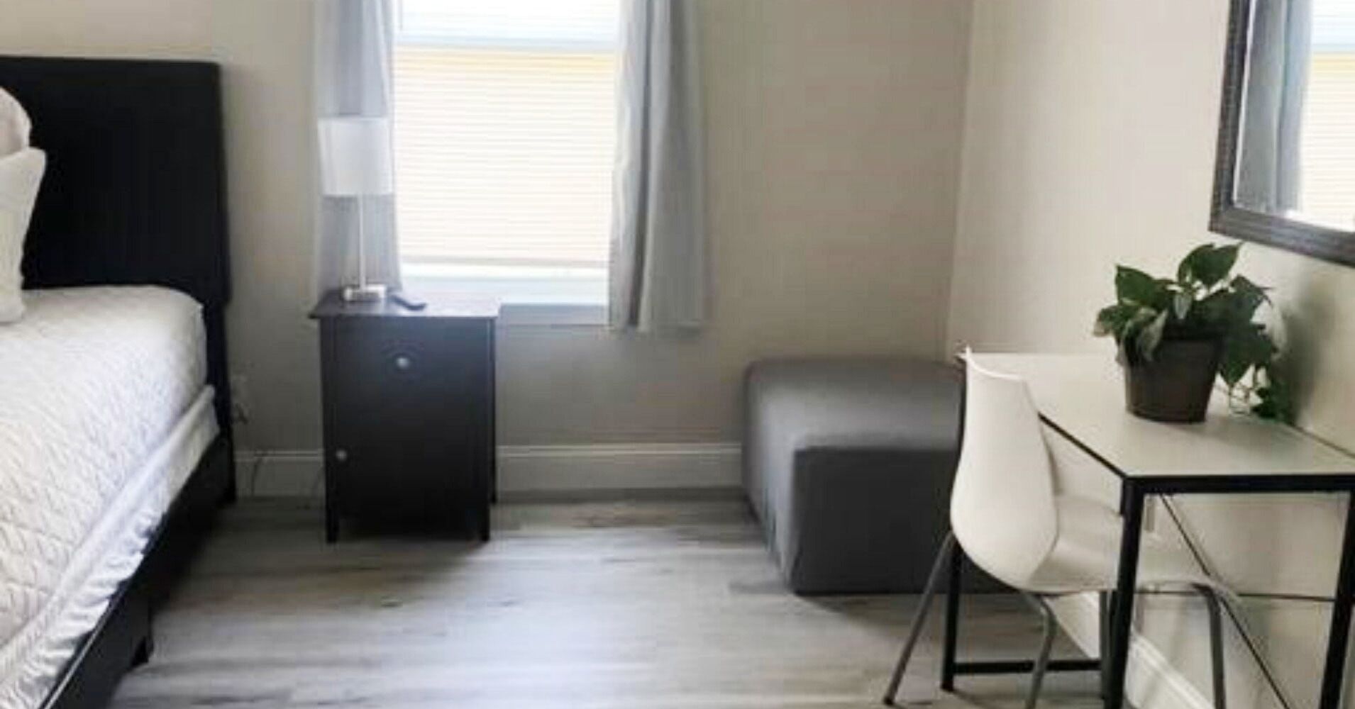 A bedroom with a bed, a nightstand, a desk, a chair, and a potted plant.