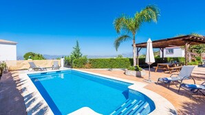 A rectangular swimming pool with clear blue water, surrounded by lounge chairs and a wooden pergola with a white umbrella.