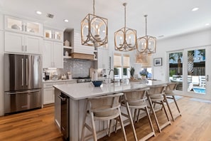A modern kitchen with white cabinetry, a stainless steel refrigerator, a large island with bar stools, and pendant lighting. - Nearly New Home w/ Heated Pool & Golf Cart, 5 min walk to Blue Mtn Beach, FL-30A (Santa Rosa Beach)