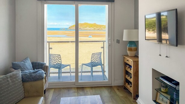 A room with a sliding glass door to a beach, a sofa, a TV, and a small wooden cabinet. - Pennti Treth - UK39455 (Porth, near Newquay)