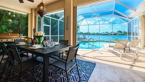 Un coin repas avec une table dressée pour quatre personnes, un ventilateur de plafond et une vue sur une piscine et un plan d’eau.