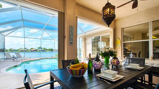 Un espace repas aménagé au bord de la piscine, avec une table dressée pour deux personnes, un saladier rempli de fruits et une bouteille de champagne.