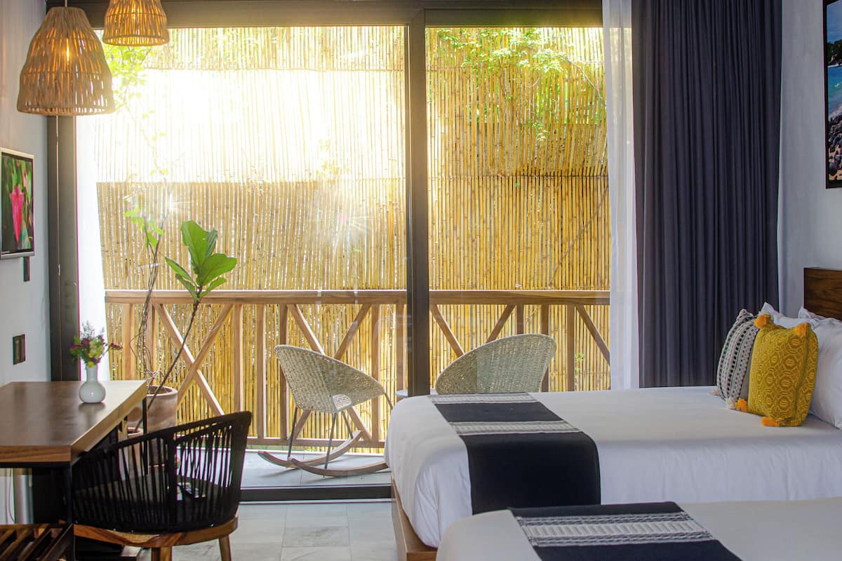 Habitación de hotel con cama, dos sillas, una mesa de comedor y un ventanal con vistas a bambú y vegetación.