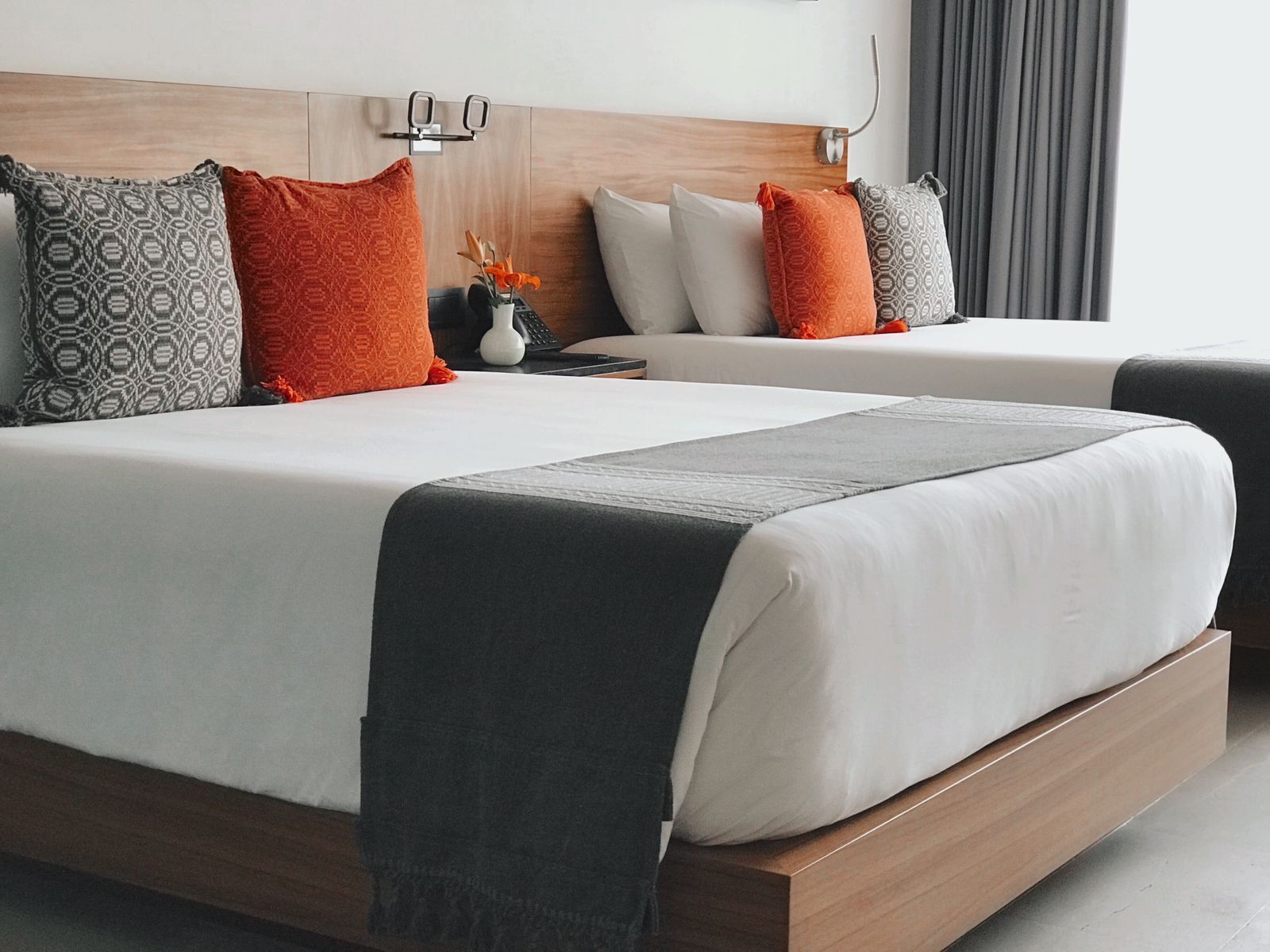 Quarto de hotel com duas camas, cada uma com roupa de cama branca e almofadas estampadas. Há uma mesa de cabeceira com um abajur e um vaso com flores.