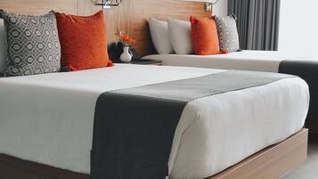 Quarto de hotel com duas camas, cada uma com roupa de cama branca e almofadas estampadas. Há uma mesa de cabeceira com um abajur e um vaso com flores.